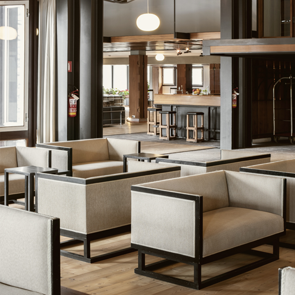 Zona lounge dell’Hotel Boite con poltrone dal design geometrico rivestite in tessuto beige e struttura in metallo nero, tavolini coordinati e pavimento in legno. Sullo sfondo si intravede il bar con sgabelli in legno e metallo, in un ambiente caldo e razionale ispirato al modernismo di Gellner.