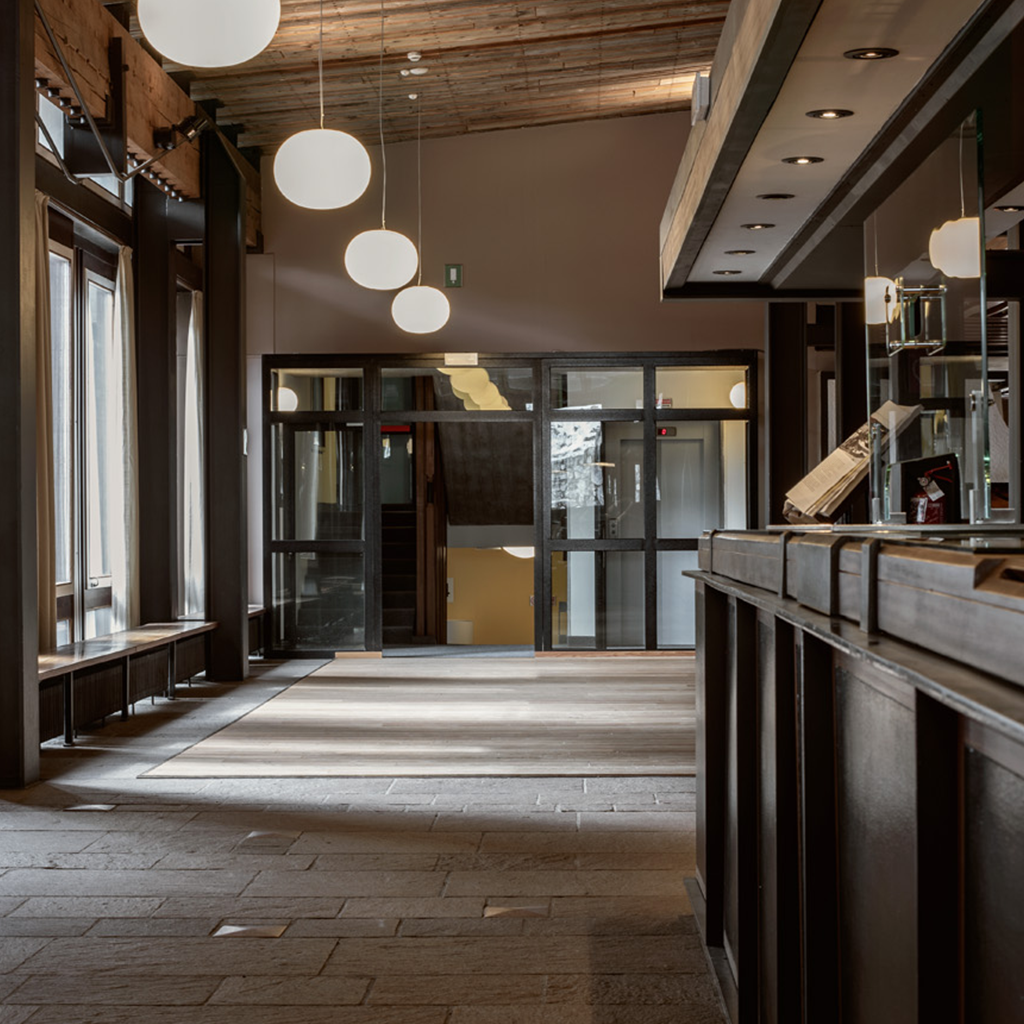 Ingresso dell’Hotel Boite con pavimentazione in pietra, luci sferiche sospese, soffitto in legno e reception in legno scuro. Le grandi vetrate offrono trasparenze e profondità, in uno spazio che coniuga accoglienza funzionale e coerenza con il progetto architettonico originario.