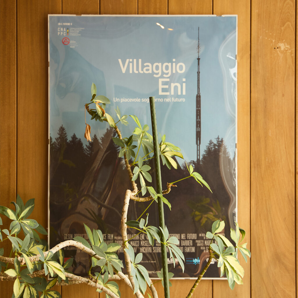 Poster of the Eni Village hanging on a wooden wall, with the slogan “A pleasant stay in the future” and an indoor plant in the foreground. A tribute to the site’s history and architectural legacy.

