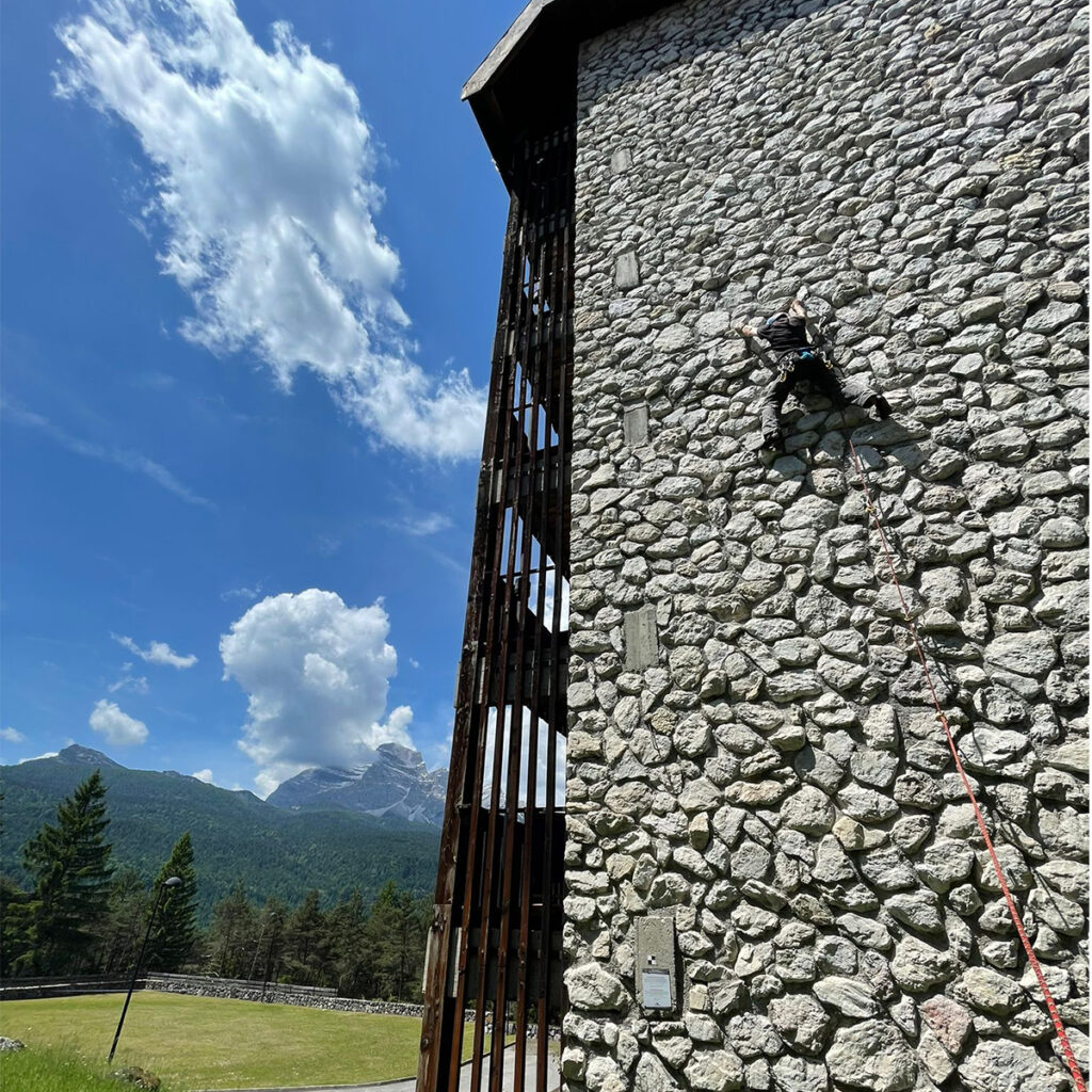 A young climber ascends the stone wall of the Hotel Boite, secured with a rope. In the background, green grass and the Dolomite mountains.