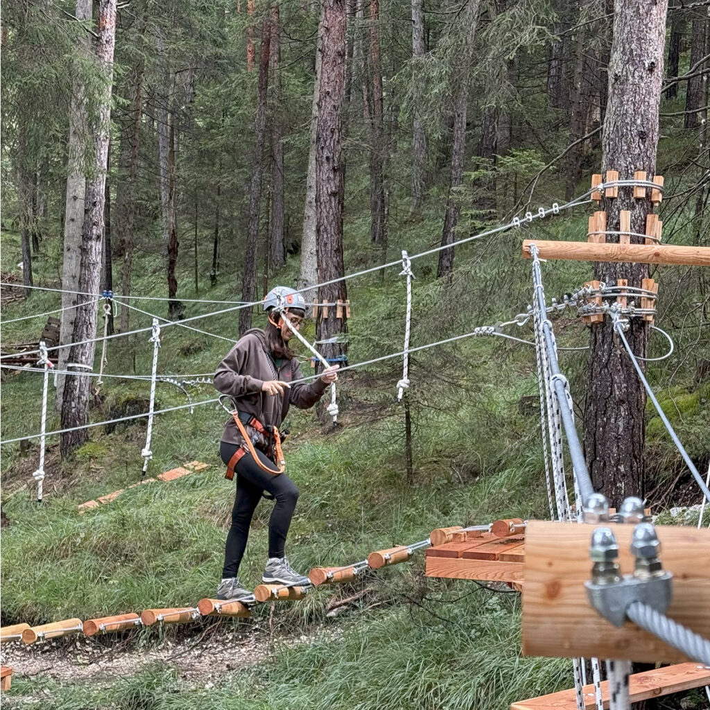 A young woman crosses the rope bridge suspended among the trees at Hotel Boite’s Adventure Park, safely equipped.