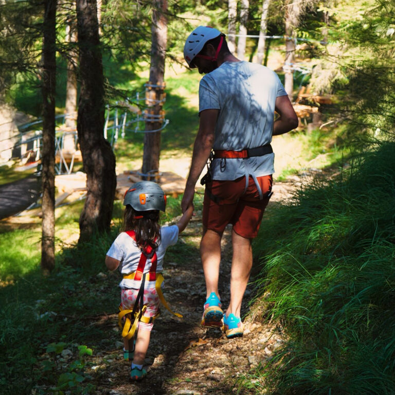 An adult and a child walk hand in hand in the forest of Hotel Boite’s Adventure Park, wearing helmets and harnesses.