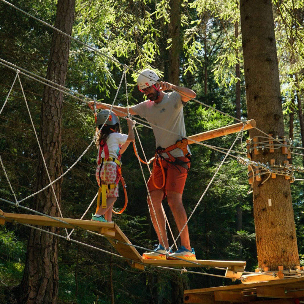 An adult guides a child across the suspension bridge at Hotel Boite’s Adventure Park, both wearing helmets and harnesses.