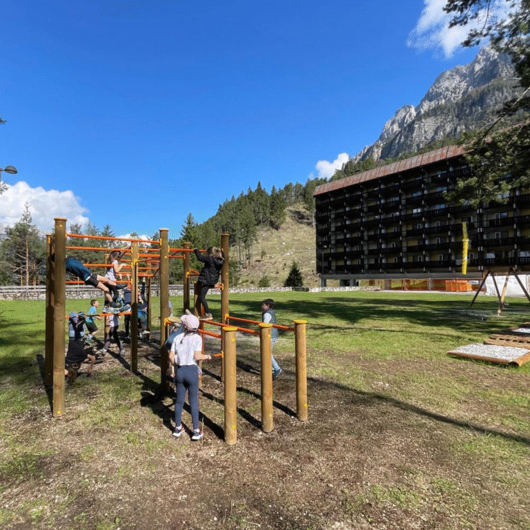 Group of children playing on a wooden and metal climbing frame in front of Hotel Boite, with the Dolomites in the background.