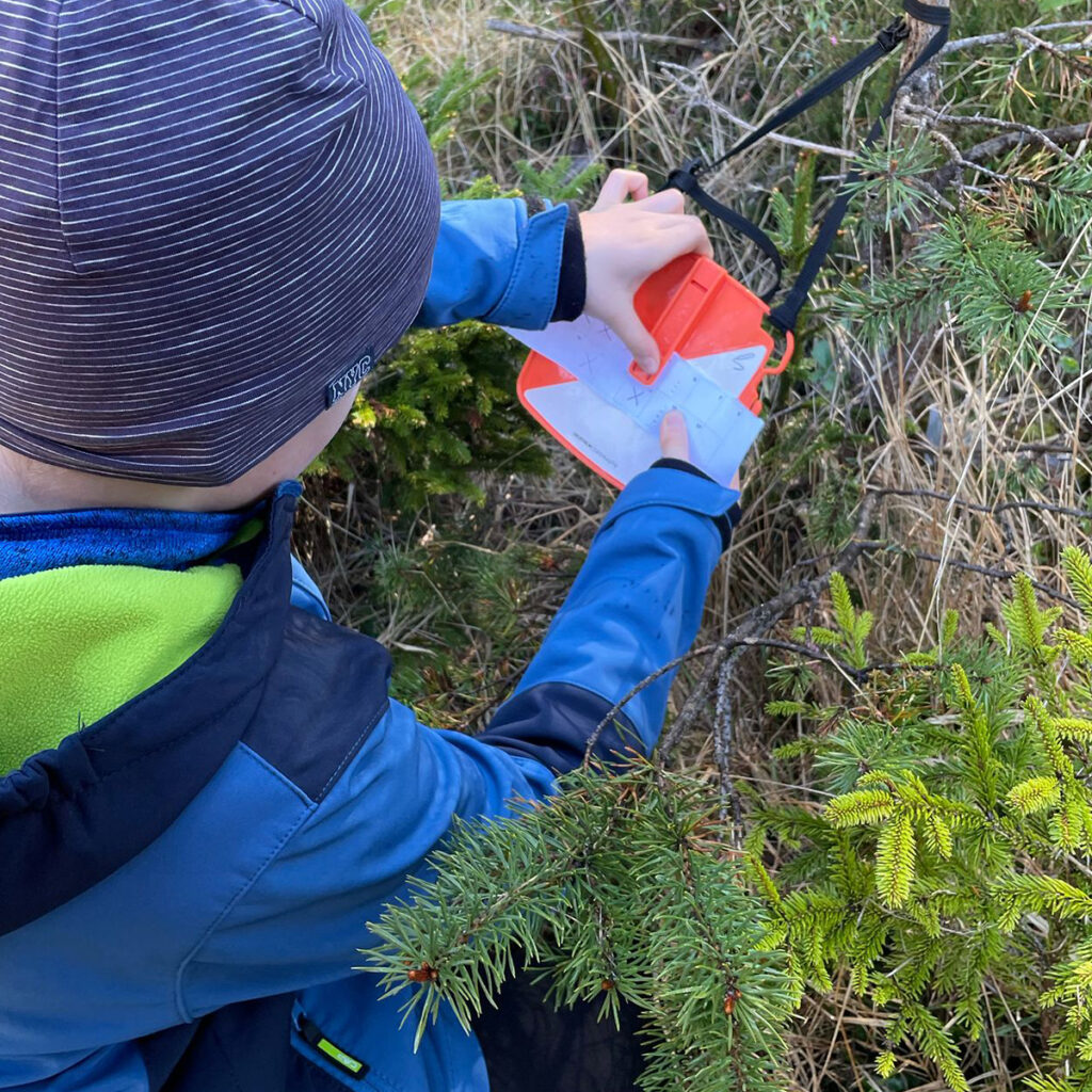 Bambino con cappellino e giacca tecnica che consulta la mappa su una lanterna da orienteering appesa a un ramo, durante un’attività educativa nei boschi attorno all’Hotel Boite.