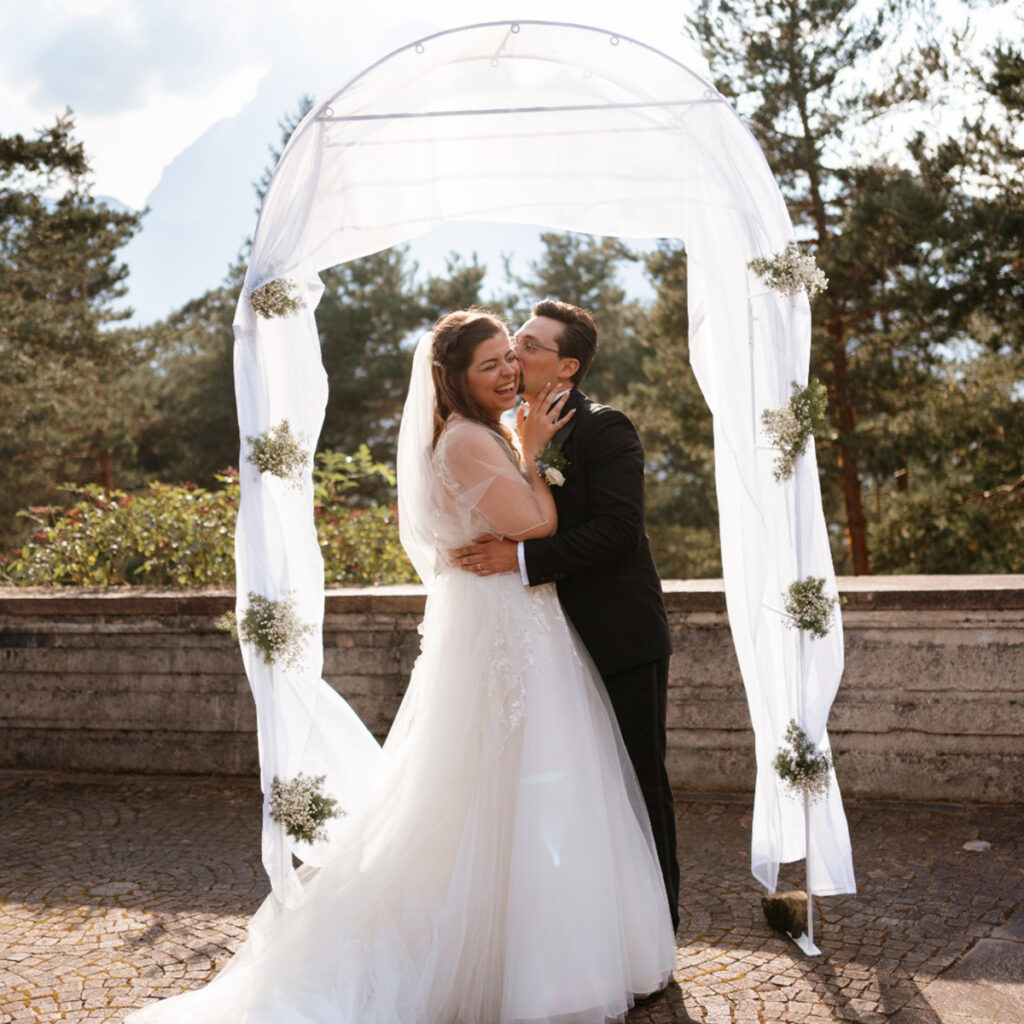 Bride and groom smile and embrace under a floral arch outdoors in the Dolomites.