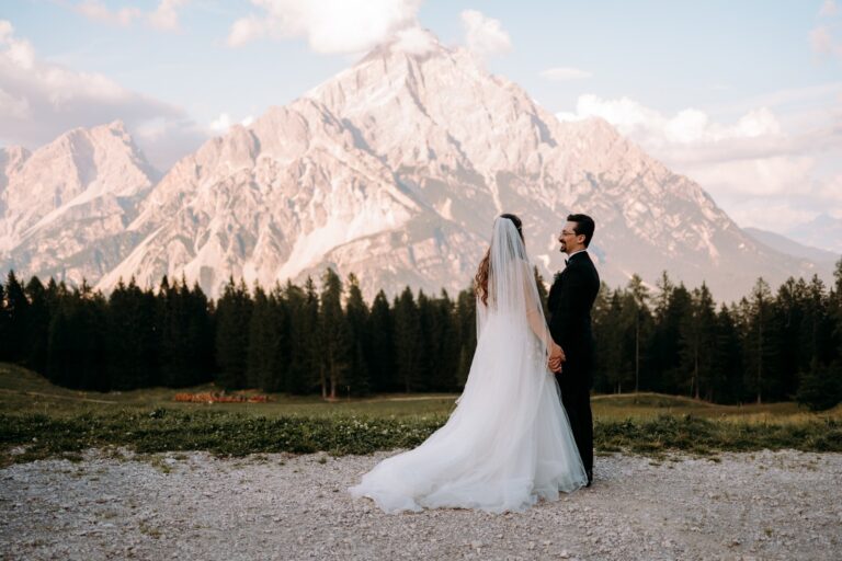A bride and groom in wedding attire hold hands facing the majestic Dolomites landscape.